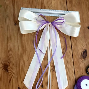 Peut inclure: Un grand nœud en satin blanc avec des rubans à queue ombrés de violet et de blanc. Le nœud est noué avec un ruban violet et se trouve sur une surface en bois. Il y a des bobines de ruban violet dans le coin inférieur droit de l'image.