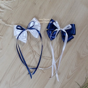 Peut inclure: Deux nœuds en ruban de satin avec de longues queues. Un nœud est blanc avec des queues bleu marine, et l'autre est bleu marine avec des queues blanches. Les nœuds sont liés ensemble avec de la ficelle.