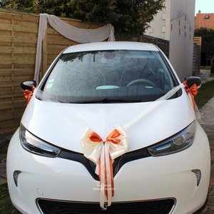 Pu&ograve; includere: Un'auto bianca decorata con un grande fiocco arancione e bianco per un matrimonio. L'auto &egrave; parcheggiata davanti a una recinzione di legno e una tenda bianca trasparente.
