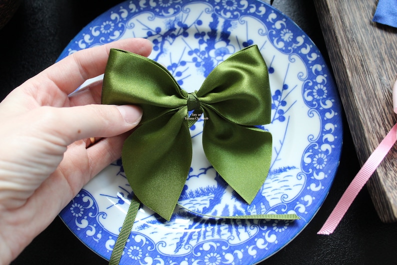 Puede incluir: Un lazo de sat&eacute;n verde oliva hecho a mano, con largas colas, se sostiene sobre un plato con estampado azul y blanco. El lazo es el punto focal, mostrando su textura suave y dise&ntilde;o sim&eacute;trico. Una cinta verde a juego se extiende desde el lazo.
