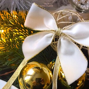 Peut inclure: Un nœud en velours blanc avec une bordure dorée, noué autour d'une boule de Noël dorée. Le nœud est noué avec un ruban doré.
