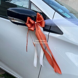 Pu&ograve; includere: Un'auto bianca decorata con un grande fiocco arancione e tulle bianco. Il fiocco &egrave; legato allo specchietto laterale dell'auto. Il testo "Idea Tatyarn Tetiana Chankrob" &egrave; stampato sulla portiera dell'auto.