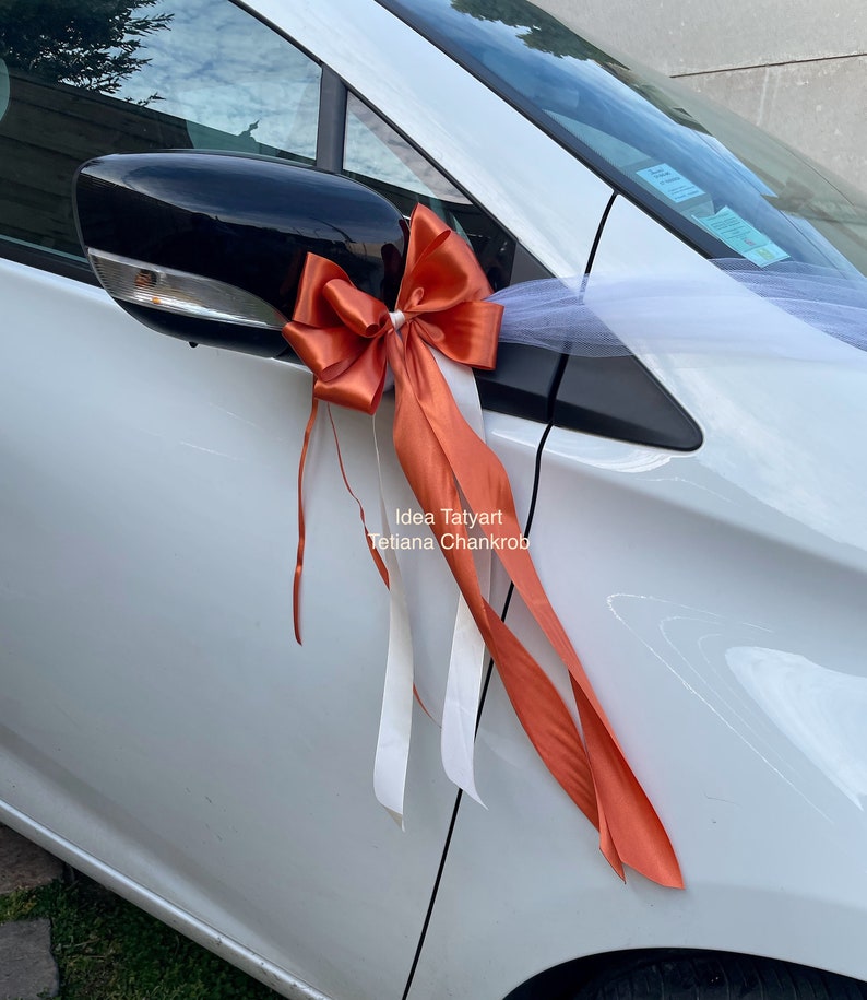 Pu&ograve; includere: Un'auto bianca decorata con un grande fiocco arancione e tulle bianco. Il fiocco &egrave; legato allo specchietto laterale dell'auto. Il testo "Idea Tatyarn Tetiana Chankrob" &egrave; stampato sulla portiera dell'auto.