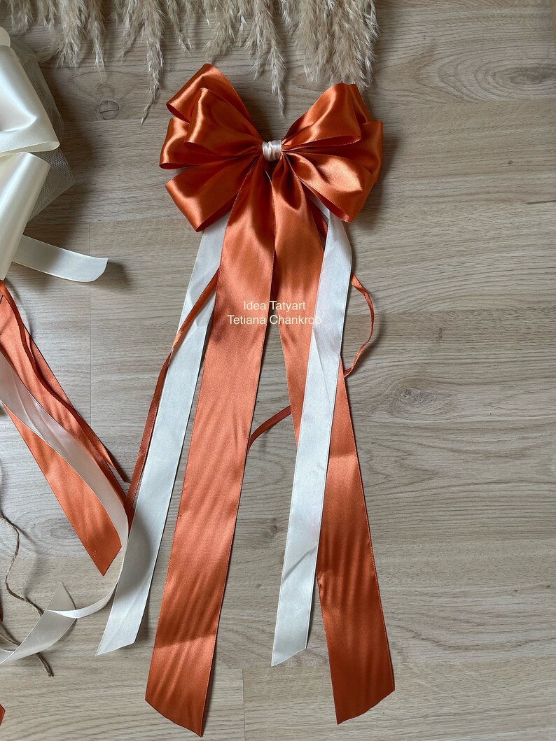 Pu&ograve; includere: Un grande fiocco di raso arancione con lunghe code di nastro di raso arancione e bianco. Il fiocco &egrave; legato con un piccolo fiocco di raso bianco al centro. Il testo "Idea Tatyarn Tetiana Chankro" &egrave; stampato sul nastro.