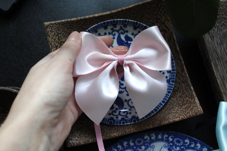 Puede incluir: Un lazo de sat&eacute;n rosa claro con largas cintas, apoyado sobre un plato con estampado azul y blanco. El lazo est&aacute; sostenido por una mano, y el fondo incluye una bandeja de madera y platos azules y blancos adicionales.