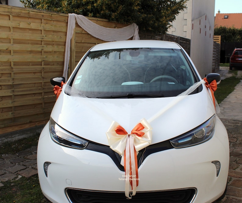 Pu&ograve; includere: Un'auto bianca decorata con un grande fiocco arancione e bianco per un matrimonio. L'auto &egrave; parcheggiata davanti a una recinzione di legno e una tenda bianca trasparente.