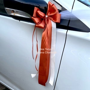 Pu&ograve; includere: Un'auto bianca decorata con un grande fiocco arancione e bianco. Il fiocco &egrave; legato allo specchietto retrovisore laterale e ha lunghi nastri che pendono. Il testo "Idea Tatyart Tatiana Chankraba" &egrave; visibile sull'auto.