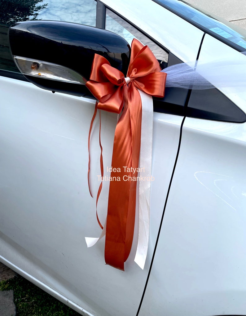 Pu&ograve; includere: Un'auto bianca decorata con un grande fiocco arancione e bianco. Il fiocco &egrave; legato allo specchietto retrovisore laterale e ha lunghi nastri che pendono. Il testo "Idea Tatyart Tatiana Chankraba" &egrave; visibile sull'auto.