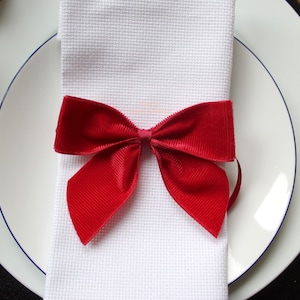 Puede incluir: Una servilleta blanca con un lazo de terciopelo rojo atado alrededor. La servilleta está sobre un plato blanco con un borde azul.
