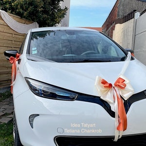 Pu&ograve; includere: Un'auto bianca decorata con un grande fiocco arancione e bianco e nastri bianchi. L'auto &egrave; parcheggiata davanti a una recinzione di legno e un muro di cemento. Il testo "Idea Tatyarn Tetiana Chankrob" &egrave; visibile sull'auto.