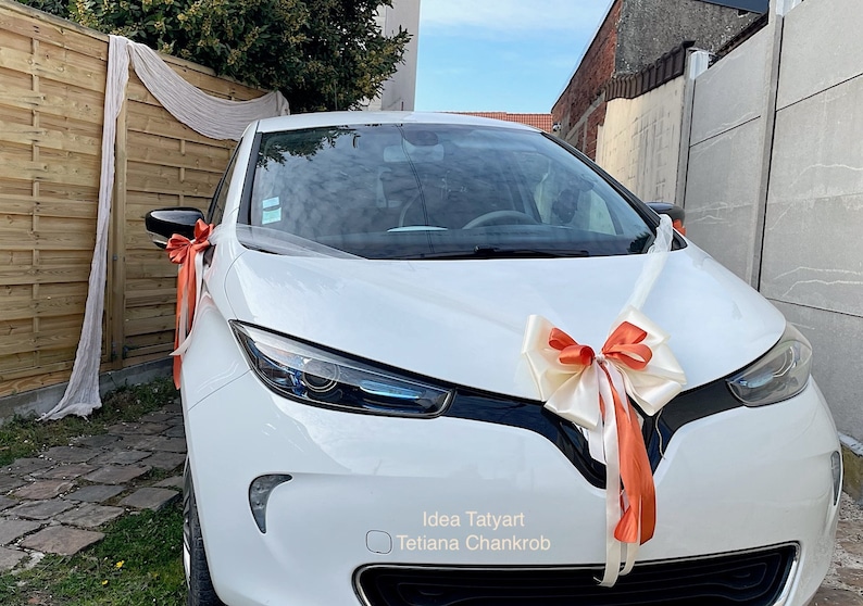 Pu&ograve; includere: Un'auto bianca decorata con un grande fiocco arancione e bianco e nastri bianchi. L'auto &egrave; parcheggiata davanti a una recinzione di legno e un muro di cemento. Il testo "Idea Tatyarn Tetiana Chankrob" &egrave; visibile sull'auto.