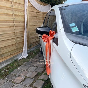 Pu&ograve; includere: Un'auto bianca decorata con un fiocco di tulle bianco e un grande fiocco arancione. L'auto &egrave; parcheggiata davanti a una recinzione di legno con una tenda bianca trasparente che pende.