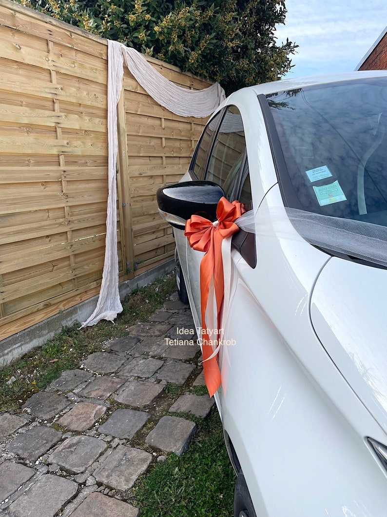 Pu&ograve; includere: Un'auto bianca decorata con un fiocco di tulle bianco e un grande fiocco arancione. L'auto &egrave; parcheggiata davanti a una recinzione di legno con una tenda bianca trasparente che pende.