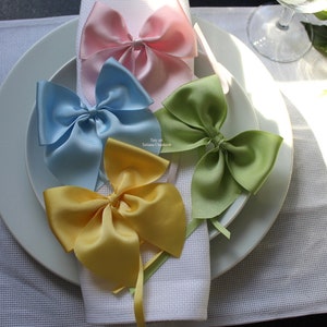 Peut inclure: Quatre nœuds en ruban de satin de différentes couleurs : rose, bleu clair, jaune et vert. Les nœuds sont disposés sur une serviette en papier blanche sur une assiette blanche. L’assiette est posée sur une nappe blanche.