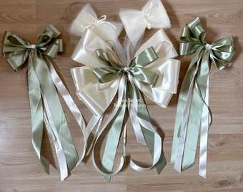 Può includere: Tre fiocchi di nastro di raso avorio e verde salvia con code lunghe. I fiocchi sono legati insieme con un tessuto di tulle.