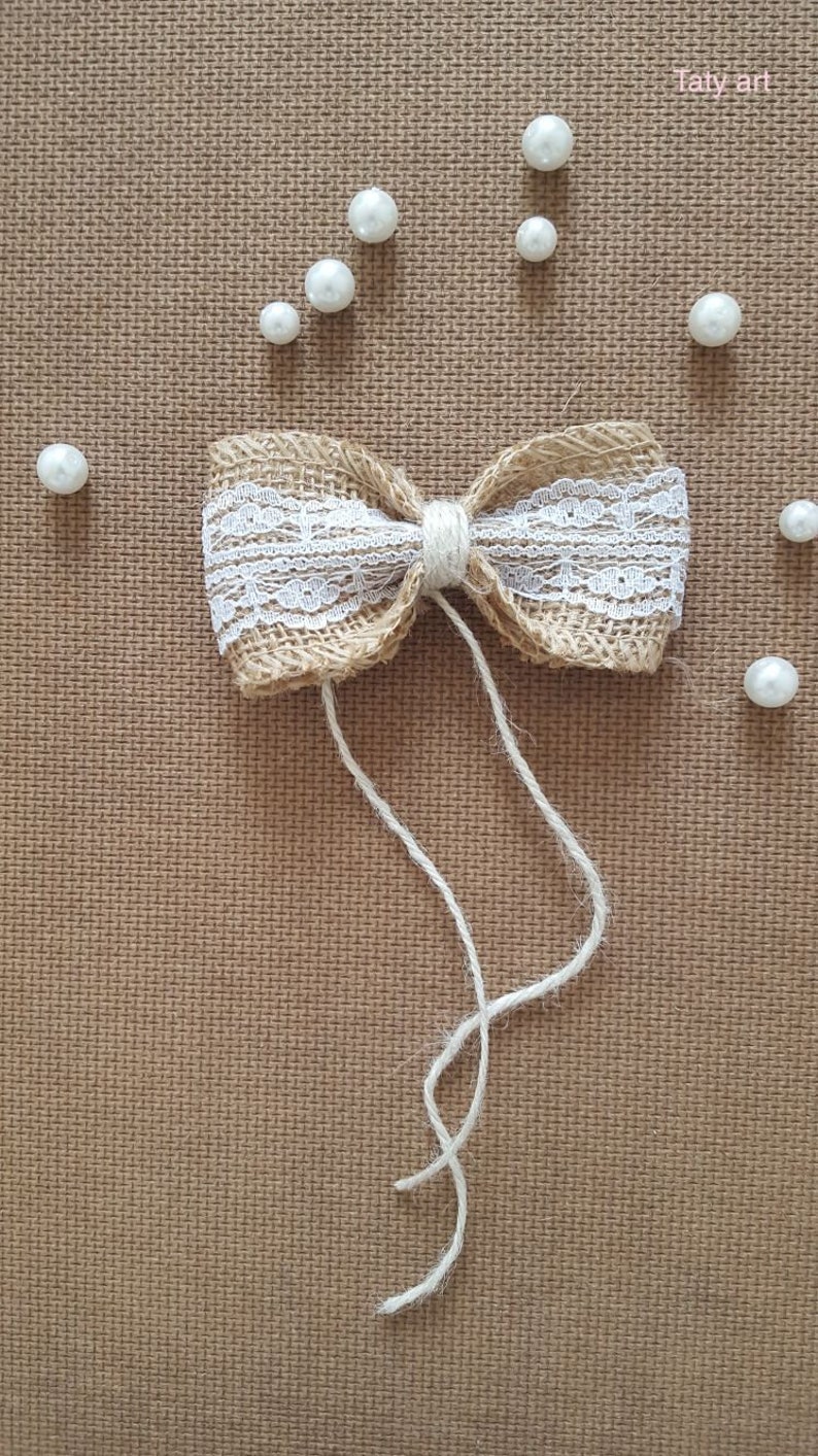 Peut inclure: Un n&oelig;ud en toile de jute marron avec une garniture en dentelle blanche et une ficelle en ficelle blanche. Le n&oelig;ud est sur un fond textur&eacute; marron avec des perles de perles blanches &eacute;parpill&eacute;es autour. Le texte "Taty art" est dans le coin sup&eacute;rieur droit de l'image.