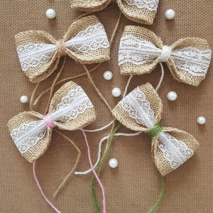 Peut inclure: Cinq n&oelig;uds en toile de jute avec une garniture en dentelle blanche et des accents de perles. Les n&oelig;uds sont attach&eacute;s avec de la ficelle et dispos&eacute;s sur un fond en toile de jute brune. Le texte "Taty art" est visible en bas de l'image.