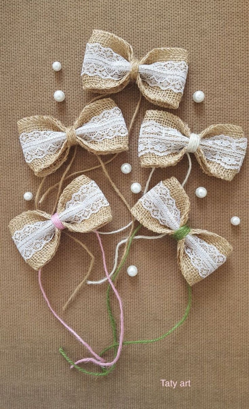 Peut inclure: Cinq n&oelig;uds en toile de jute avec une garniture en dentelle blanche et des accents de perles. Les n&oelig;uds sont attach&eacute;s avec de la ficelle et dispos&eacute;s sur un fond en toile de jute brune. Le texte "Taty art" est visible en bas de l'image.
