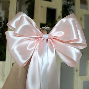 Peut inclure: Un grand nœud en satin rose clair avec un ruban blanc. Le nœud est tenu devant un fond en bois. Le nœud a plusieurs couches et une finition brillante. Le texte "Idea Tatyart, Tetiana Charkrob" est visible sur le nœud.