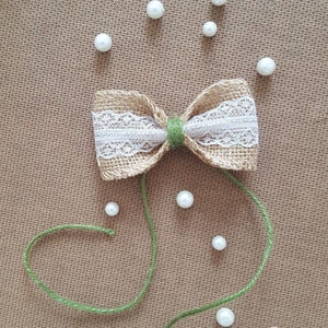 Peut inclure: Un n&oelig;ud en toile de jute avec une garniture en dentelle blanche et un ruban vert. Le n&oelig;ud est entour&eacute; de perles de perles blanches sur une surface textur&eacute;e brune. Le texte "Taty art" est dans le coin inf&eacute;rieur droit de l'image.