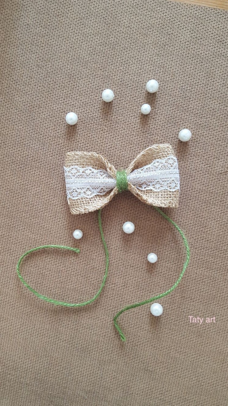 Peut inclure: Un n&oelig;ud en toile de jute avec une garniture en dentelle blanche et un ruban vert. Le n&oelig;ud est entour&eacute; de perles de perles blanches sur une surface textur&eacute;e brune. Le texte "Taty art" est dans le coin inf&eacute;rieur droit de l'image.