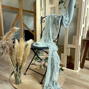 Peut inclure: Un tissu léger bleu clair drapé sur une chaise en métal noir avec un siège en bois. La chaise est sur une plate-forme ronde en bois. Il y a des herbes de pampa séchées dans un vase en verre sur la plate-forme. L'arrière-plan est un mur en bois avec un motif de grille.
