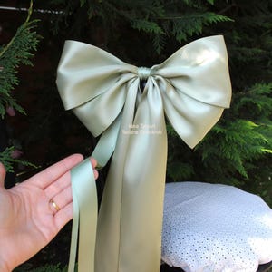 Op de afbeelding: Een grote satijnen strik in een zachte, gedempte groene kleur. De strik heeft lange, vloeiende linten en is in het midden geknoopt. De afbeelding toont ook een hand die een van de linten vasthoudt. De strik is waarschijnlijk voor decoratie of cadeauverpakking.