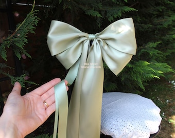 May include: A large, satin bow in a soft, muted green color. The bow has long, flowing ribbons and is tied in the center. The image also shows a hand holding one of the ribbons. The bow is likely for decoration or gift wrapping.