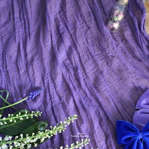 Puede incluir: Tela morada con superficie texturizada. La tela está drapeada sobre una superficie de madera. Hay flores y una pequeña botella de vidrio con flores secas.