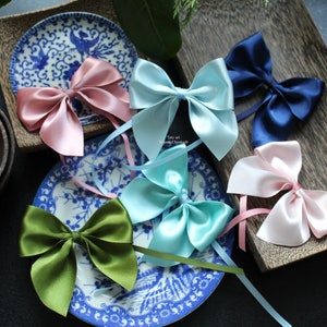 Puede incluir: Surtido de lazos de sat&eacute;n en varios colores, incluyendo rosa, azul y verde. Los lazos est&aacute;n dispuestos sobre platos decorativos azules y blancos y una bandeja de madera, ideal para manualidades o envolver regalos.