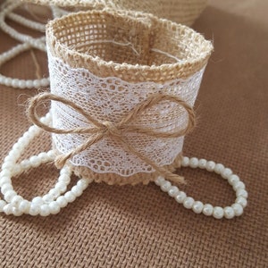 May include: A brown burlap napkin ring with a white lace trim and a brown burlap bow. The ring is surrounded by a string of white pearls.