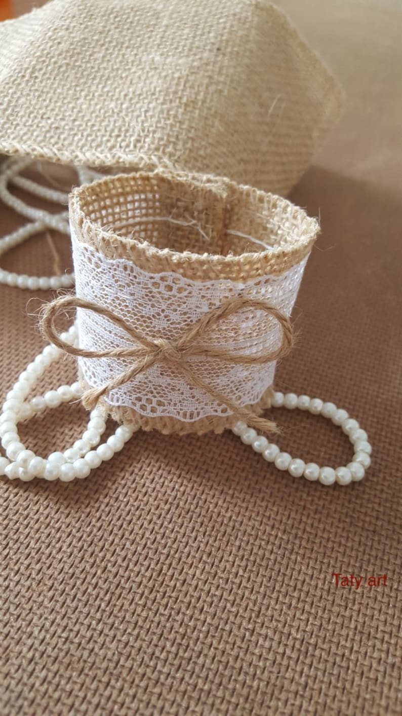 May include: A brown burlap napkin ring with a white lace trim and a brown burlap bow. The ring is surrounded by a string of white pearls.