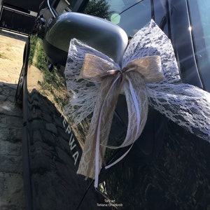 Puede incluir: Un coche negro decorado con un lazo de encaje blanco y arpillera para una boda. El lazo está atado alrededor del espejo lateral del coche.
