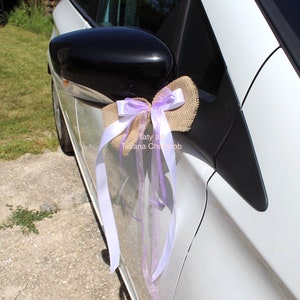 Puede incluir: Un coche blanco decorado con un lazo de arpillera y cintas de color lavanda. El lazo está atado al espejo retrovisor lateral del coche.