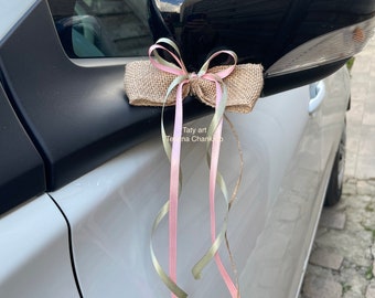 Puede incluir: Un coche blanco con un lazo de arpillera y cinta atado al espejo lateral. El lazo está decorado con cintas rosadas y verdes.