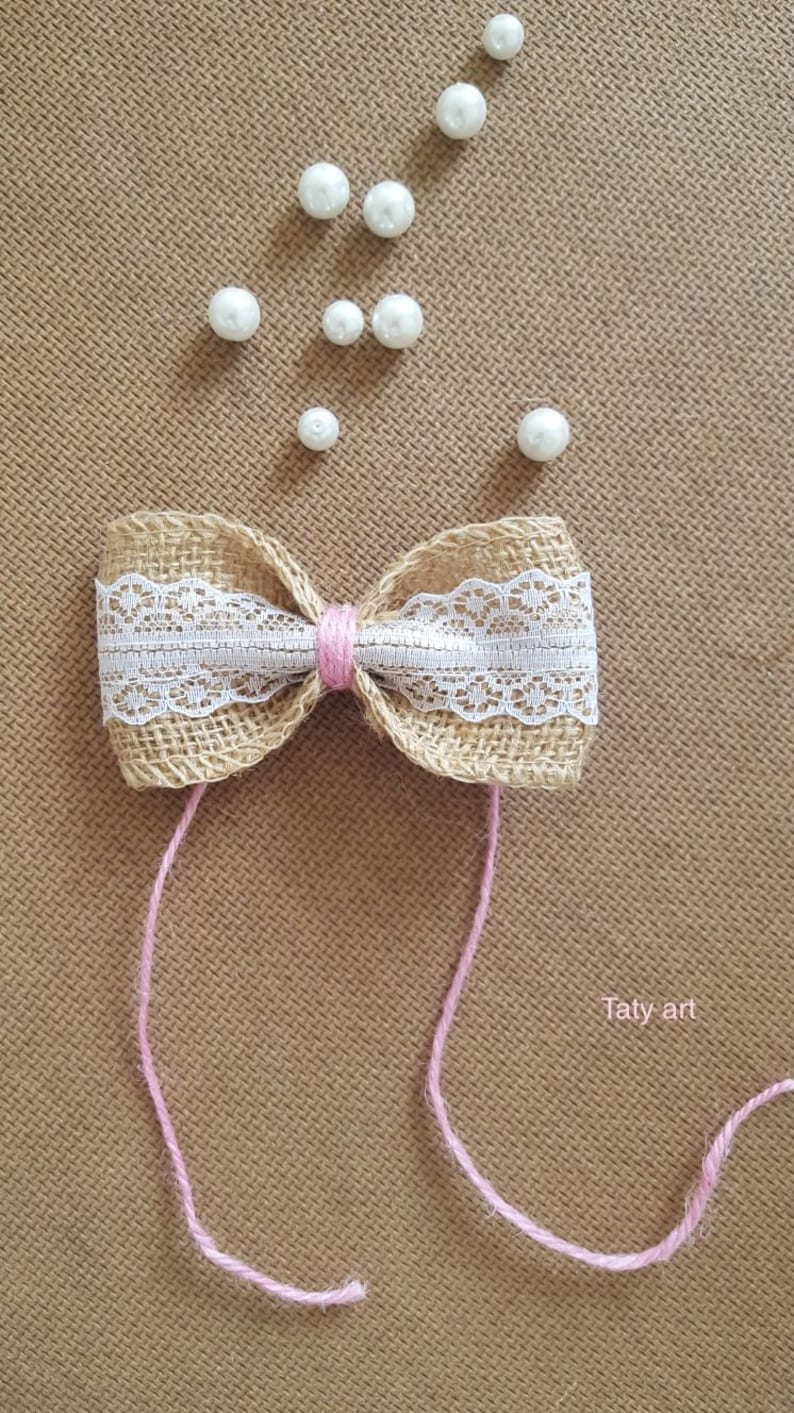 Peut inclure: Un n&oelig;ud en toile de jute avec une bordure en dentelle blanche et un ruban rose nou&eacute; au centre. Le n&oelig;ud est entour&eacute; de perles blanches sur un fond brun. Le texte "Taty art" est visible dans le coin inf&eacute;rieur droit.