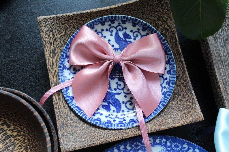 Puede incluir: Un lazo de sat&eacute;n rosa p&aacute;lido se encuentra sobre un plato con un dise&ntilde;o azul y blanco. El plato est&aacute; sobre un cuadrado de madera. El lazo tiene largas cintas. El texto "Taty art Tatiana Chankova" es visible.