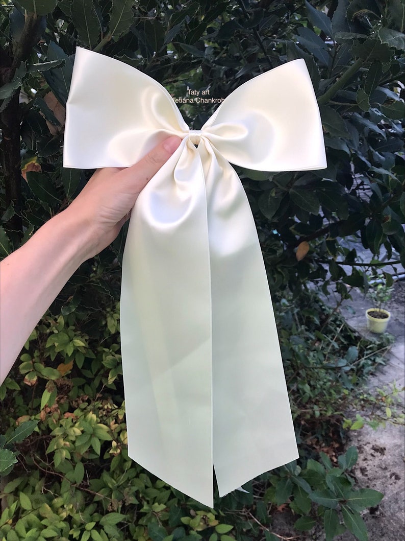 May include: A large, ivory satin bow with long tails. The bow is tied in a knot and the tails are hanging down.