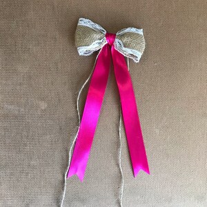 May include: A pink satin ribbon tied into a bow with a burlap and lace overlay. The bow is tied with twine and the ends of the ribbon are trimmed into points.