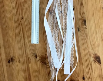 May include: White satin ribbon with lace trim tied into a bow with long tails. The ribbon is on a wooden surface.
