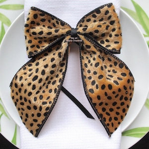 Peut inclure: Un nœud papillon en imprimé léopard brun et noir avec une bordure noire, noué sur une serviette en papier blanche, sur une assiette blanche avec des motifs de feuilles vertes.