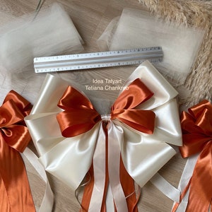 Pu&ograve; includere: Due grandi fiocchi di raso color crema e arancio bruciato con code lunghe. I fiocchi sono legati insieme con un fiocco pi&ugrave; piccolo al centro. I fiocchi sono su una superficie di legno con due pezzi di tulle e un righello.