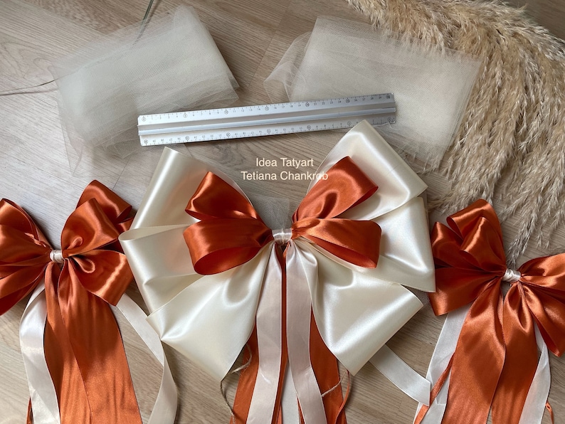Pu&ograve; includere: Due grandi fiocchi di raso color crema e arancio bruciato con code lunghe. I fiocchi sono legati insieme con un fiocco pi&ugrave; piccolo al centro. I fiocchi sono su una superficie di legno con due pezzi di tulle e un righello.