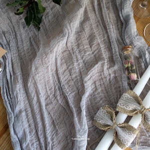 Puede incluir: Dos velas blancas con lazos de arpillera y encaje sobre un mantel de lino gris. Una pequeña botella de vidrio con flores secas está sobre la mesa. El texto "Hello Vintage" está escrito en una tarjeta marrón.