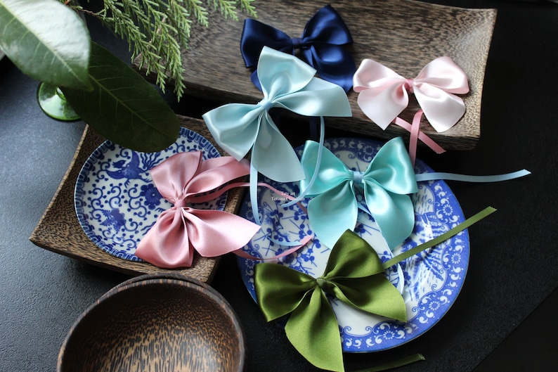 May include: Assortment of six satin ribbon bows in various colours, including light blue, pink, green, and navy blue. The bows are displayed on a blue and white patterned plate and a wooden tray.