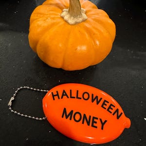 May include: An orange pumpkin with a beige stem sits above an orange oval tag. The tag reads "HALLOWEEN MONEY" in black capital letters. A silver chain is attached to the tag.
