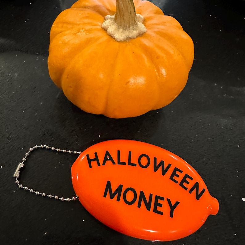 May include: An orange pumpkin with a beige stem sits above an orange oval tag. The tag reads "HALLOWEEN MONEY" in black capital letters. A silver chain is attached to the tag.