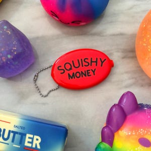 Könnte beinhalten: Eine rote, ovale Geldbörse mit dem weißen Schriftzug "SQUISHY MONEY". Sie ist an einer silbernen Kette befestigt. Im Hintergrund befinden sich weitere bunte, quetschbare Spielzeuge, darunter ein lila, blaues und oranges.