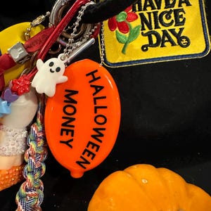 May include: A collection of keychains and charms, including a white ghost, an orange Halloween-themed coin purse with the words "Halloween Money", and a yellow patch that says "Have a Nice Day".