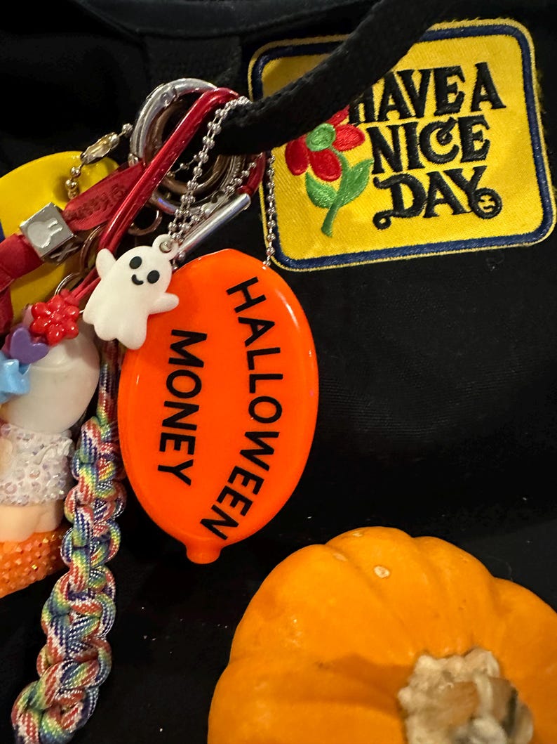 May include: A collection of keychains and charms, including a white ghost, an orange Halloween-themed coin purse with the words "Halloween Money", and a yellow patch that says "Have a Nice Day".